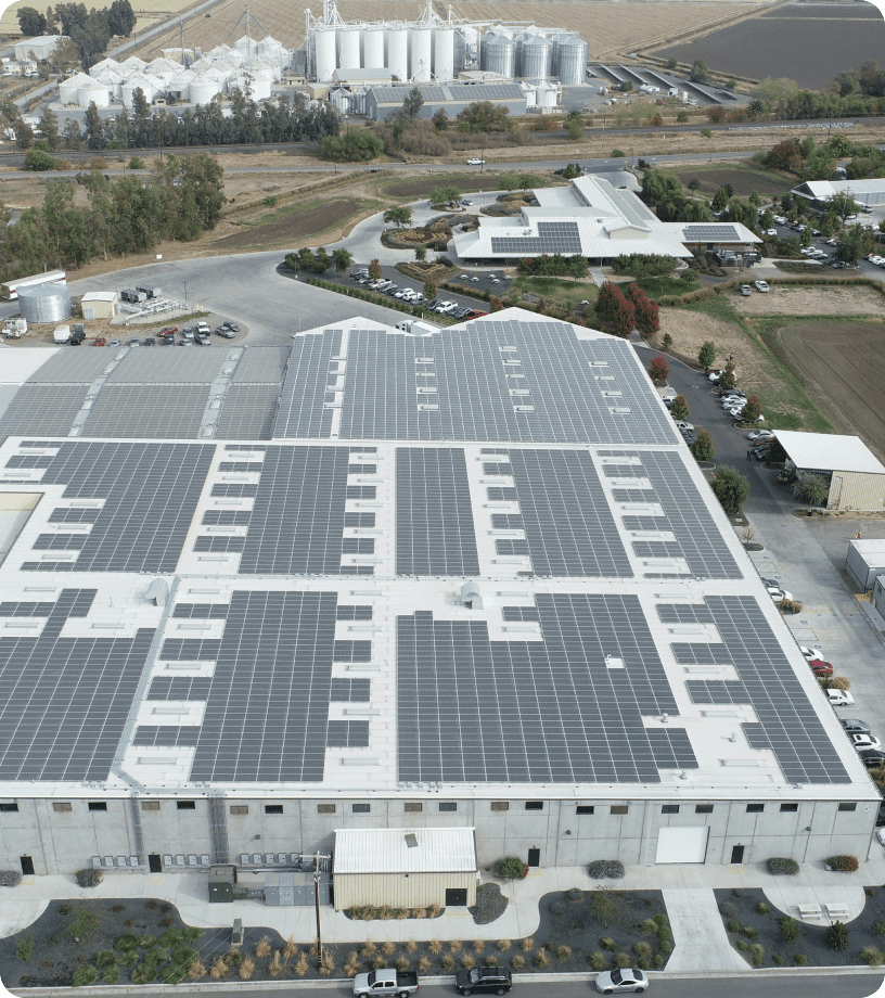 Aerial view of an industrial complex equipped with extensive rooftop solar panels installed by a local solar company, surrounded by fields, roads, and additional buildings. In the background, large storage silos and agricultural land are visible.