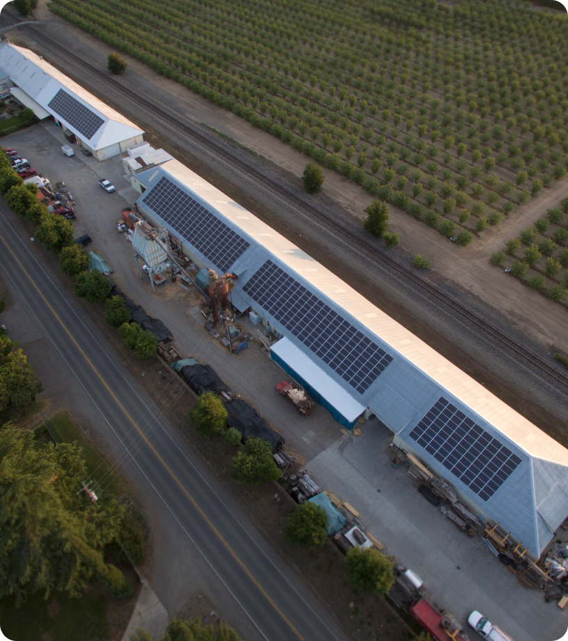 Aerial view of a large facility with multiple warehouses featuring Solar PV Systems on their roofs, surrounded by farmland and adjacent to a road and railway track.