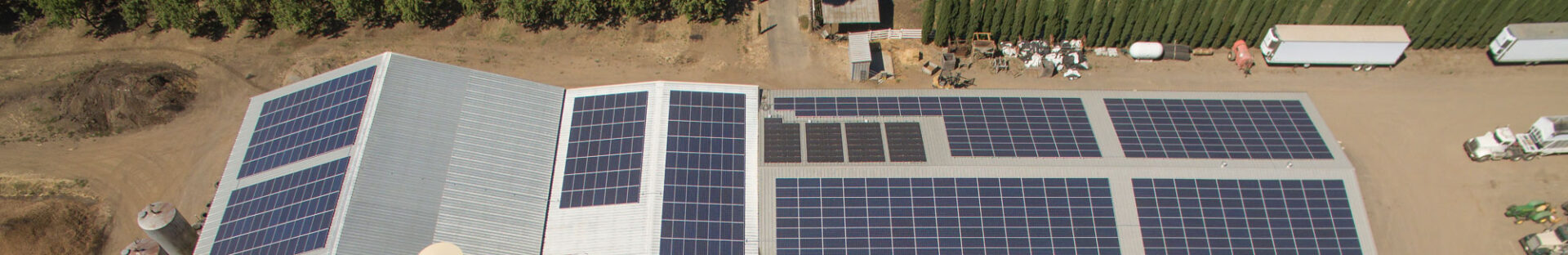 Aerial view of a large building with multiple solar panels installed on its roof, funded by a USDA REAP Grant, surrounded by trees and scattered vehicles.