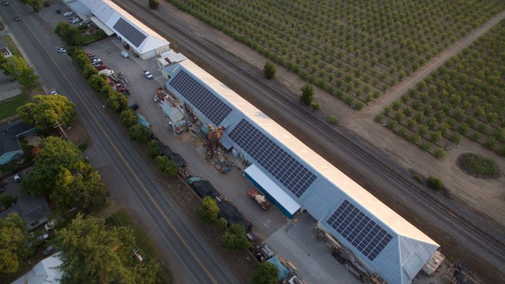 Aerial view of two long industrial buildings with solar panels on their roofs beside a road and adjacent to a large field with rows of crops, funded by a USDA REAP Grant. Vehicles are parked along the road.