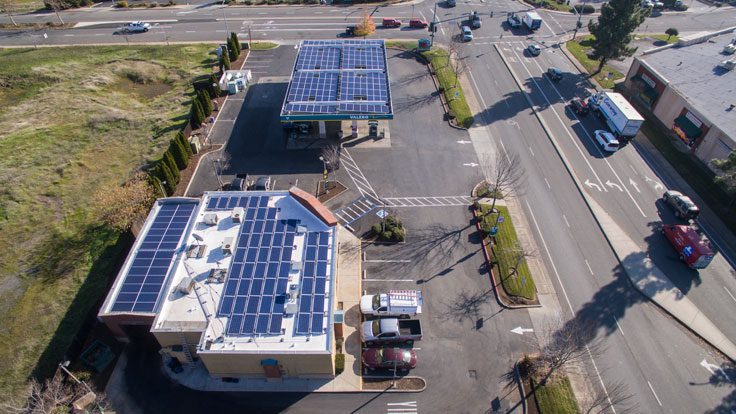 Aerial view of a small commercial area with two buildings equipped with commercial solar panels on their roofs, adjacent parking spaces, and surrounding streets with various vehicles.