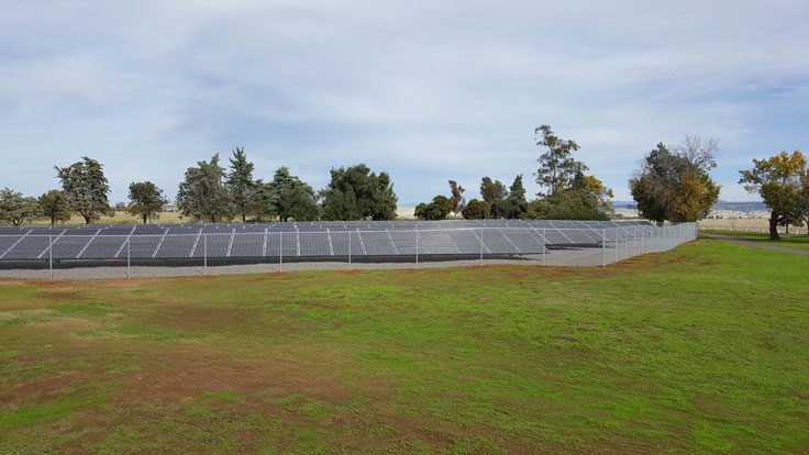 A fenced solar farm with numerous commercial solar panels is situated on a grassy field with trees in the background under a cloudy sky.