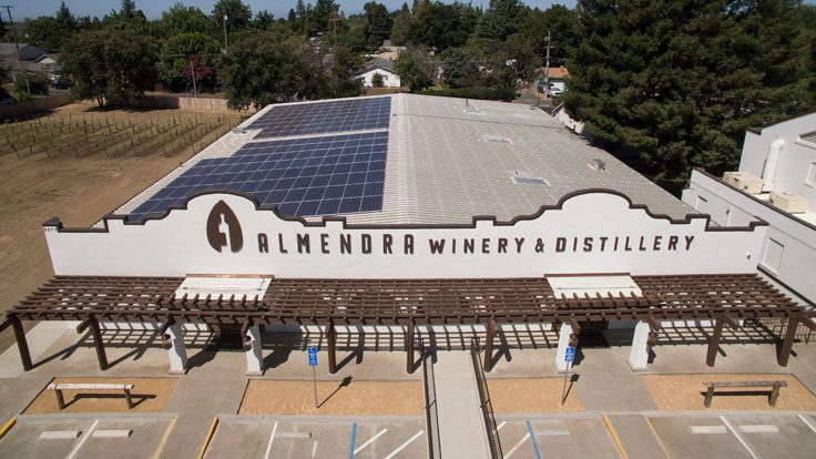 Aerial view of Almendra Winery & Distillery building with commercial solar panels on the roof and signage on the front. The surrounding area includes greenery and a small vineyard.