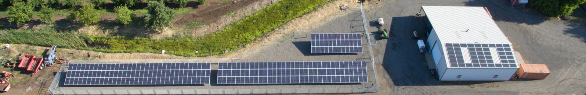 Aerial view of a facility with three solar panel arrays on land and several on the roof of a nearby building, surrounded by vegetation and machinery. This efficient layout showcases the expertise of project management consulting in optimizing space and resources.