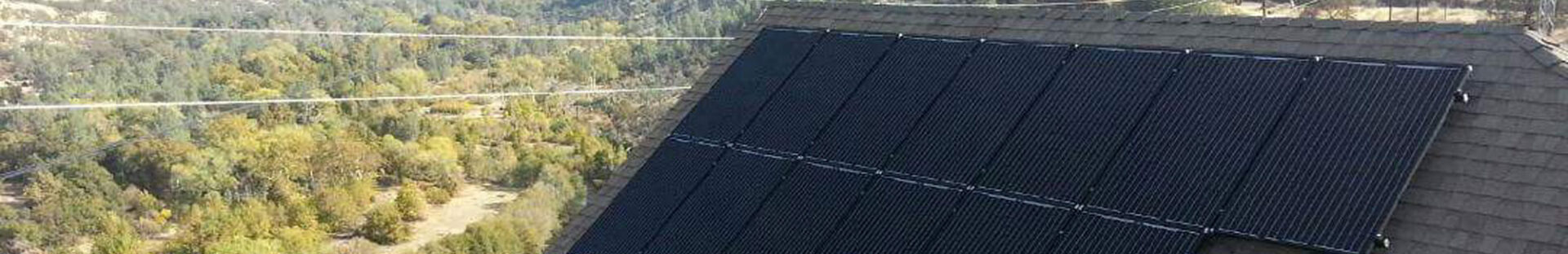 Residential solar panels installed on a shingled roof with a forested landscape in the background. Power lines are visible above the trees.