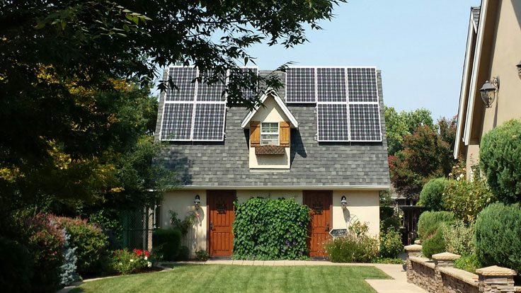 A residential house with solar panels on the roof, surrounded by a well-maintained lawn and garden with various trees and plants.