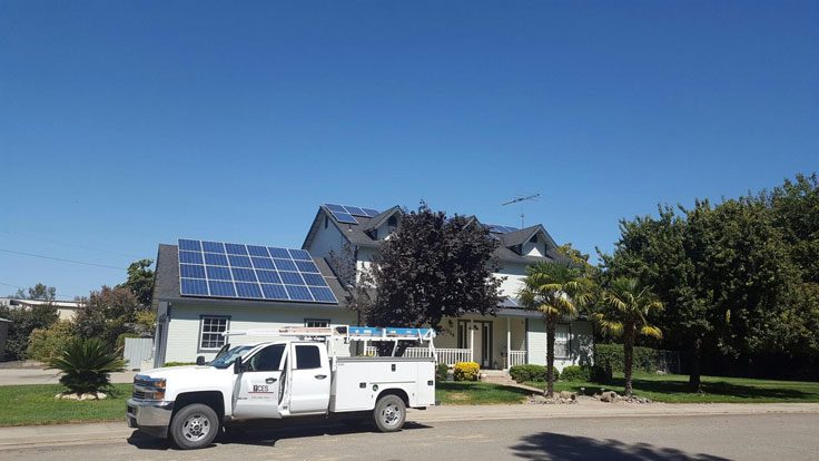 A white service truck is parked on a street in front of a house equipped with residential solar panels on the roof, under a clear blue sky.