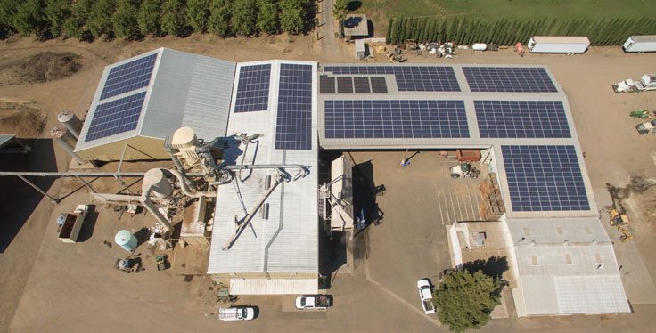 An aerial view of a facility with multiple buildings, all with rooftops covered in Solar PV Systems, surrounded by trees, vehicles, and equipment.