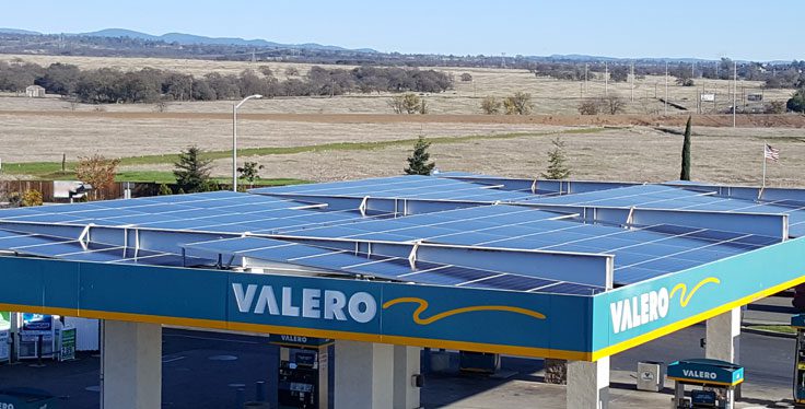 A view of a Valero gas station with Solar PV Systems on its roof, set against a backdrop of flat, open countryside and distant low hills.